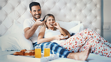 Couple laying in bed enjoying breakfast while watching TV.